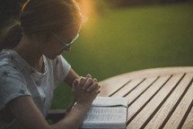 a young woman praying over an open Bible outdoors 