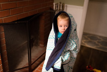 a toddler boy standing by a fireplace wrapped in a blanket 