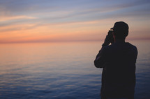 man taking a picture of the ocean at sunset 