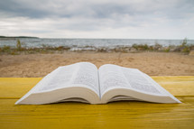 pages of an open Bible on a yellow table with beach in background 