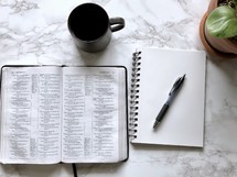 house plant, open Bible, pen, notebook, and coffee cup on a marble countertop 