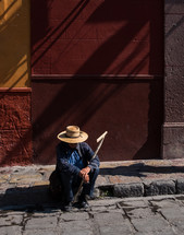 an elderly man with a walking stick resting on a curb 