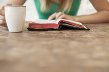 Woman reading the Bible on a wood table with a coffee cup.