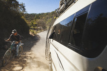 man riding a bicycle beside of a van on a dirt road 