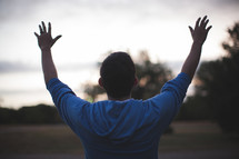 Back of man with arms raised in praise standing outside.