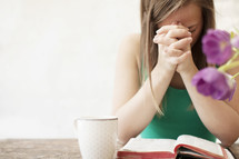 Woman with a coffee cup praying in the morning over the Bible on a wood table with flowers.