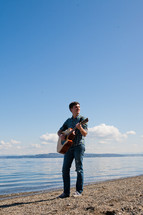 man standing in front of a lake playing a guitar 