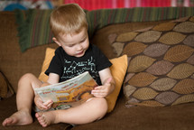 a toddler boy sitting on a couch reading a book 