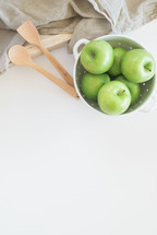 A colander of green apples, wooden utensils and a kitchen towel on a white surface.