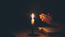 Woman's hands praying on Holy Bible with candlelight.