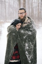 a couple hugging under a blanket in the snow 
