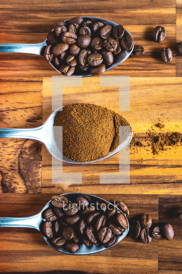 Spoons with coffee powder and coffee beans, laying down on a wooden background.