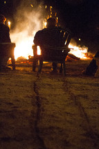 Man drags a Muskoka chair across a beach towards a large bonfire.