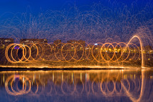 Hot Golden Sparks Flying from Man Spinning Burning Steel Wool near River with Water Reflection. Long Exposure Photography using Steel Wool Burning