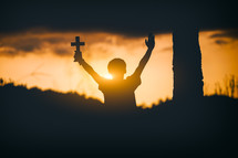 silhouette of a young man holding a cross in worship at sunset 