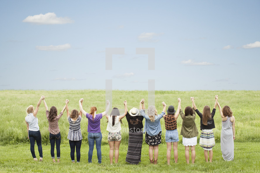 Women holding hands with arms raised in praise while standing in a field of grass.