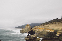 a man sitting on a rock along a shore looking out at the ocean 