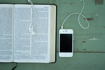 Iphone and earbuds on a wooden table with an open Bible.