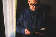 an elderly man reading a Bible standing by a window 