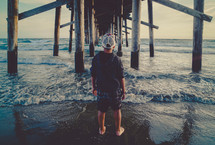 Young man standing under a pier | Contemplation | Watching Waves | Facing Forward | Standing | Back | Outside | Looking Out | Vision | Perspective | Landscape | Nature | Creation | Person | Male | Youth | Generation | Reflection