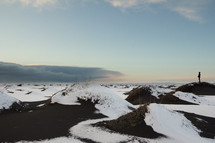 Dark clouds build in the distance over snow covered black sand dunes near Vik, Iceland on the south coast of the island. A person is standing on one of the dunes watching the approaching storm.