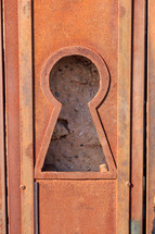 Large keyhole in a rusted metal door with a view of the rock wall behind
