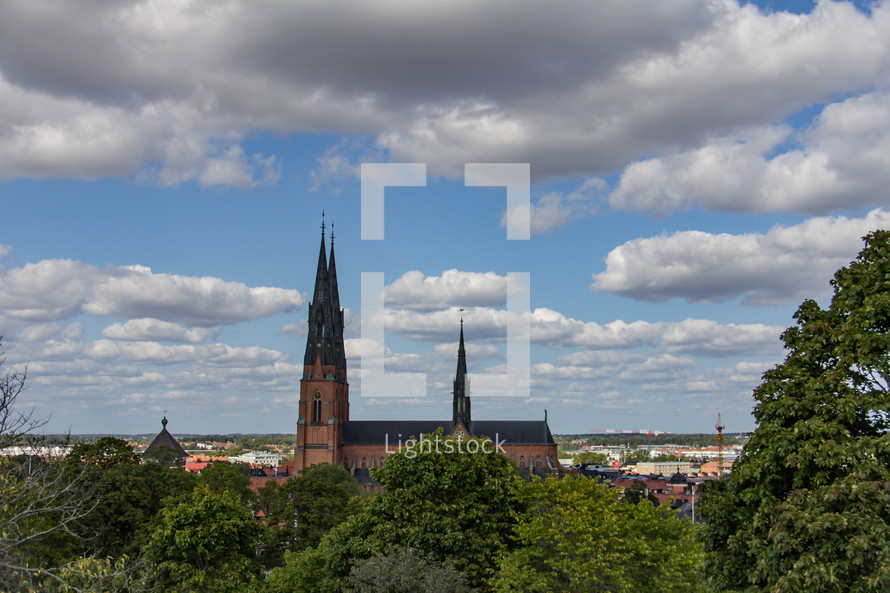 Profile of Uppsala Cathedral in Sweden under a blue sky with fluffy white clouds, green trees in the foreground