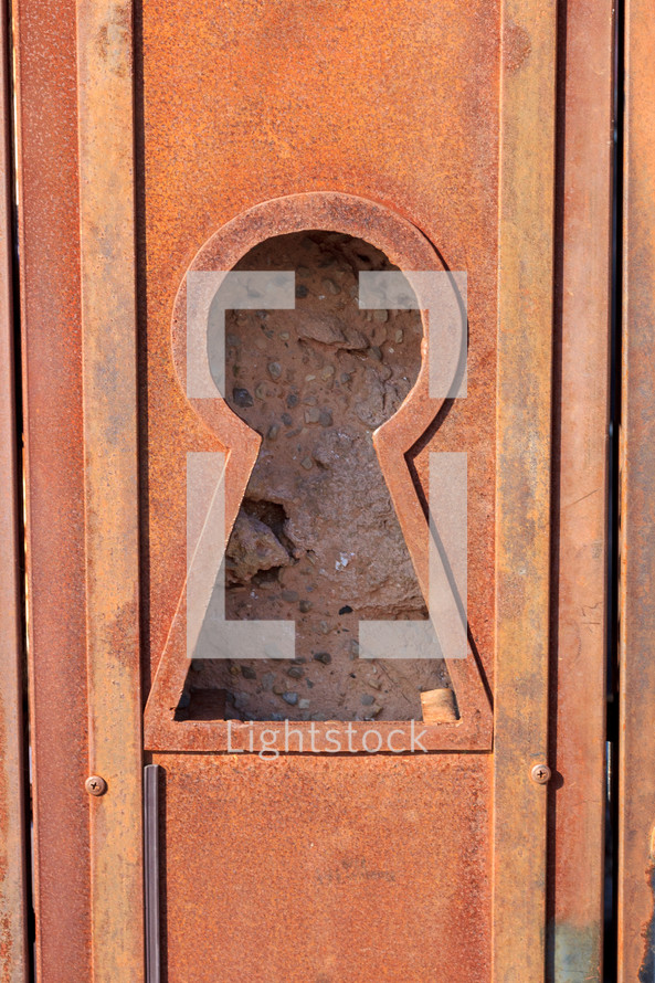 Large keyhole in a rusted metal door with a view of the rock wall behind