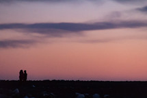 Two people walking across a rocky landscape in silhouette against a colorful sky at sunset