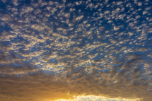 Textured clouds reflect the yellow and orange light of sunset against a blue sky