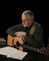 man with his arm resting on a guitar reading sheet music 