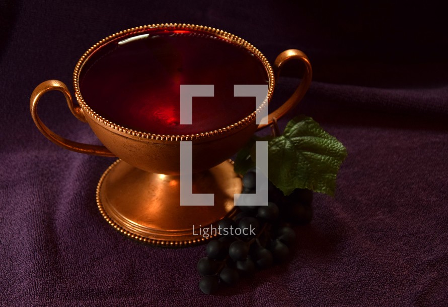 Golden colored chalice with Red liquid and grapes next to it on top of a purple cloth
