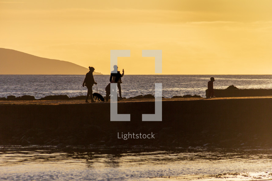 A family walking on a Salthill, Galway, Ireland jetty at sunset appear in silhouette against a yellow sky at sunset