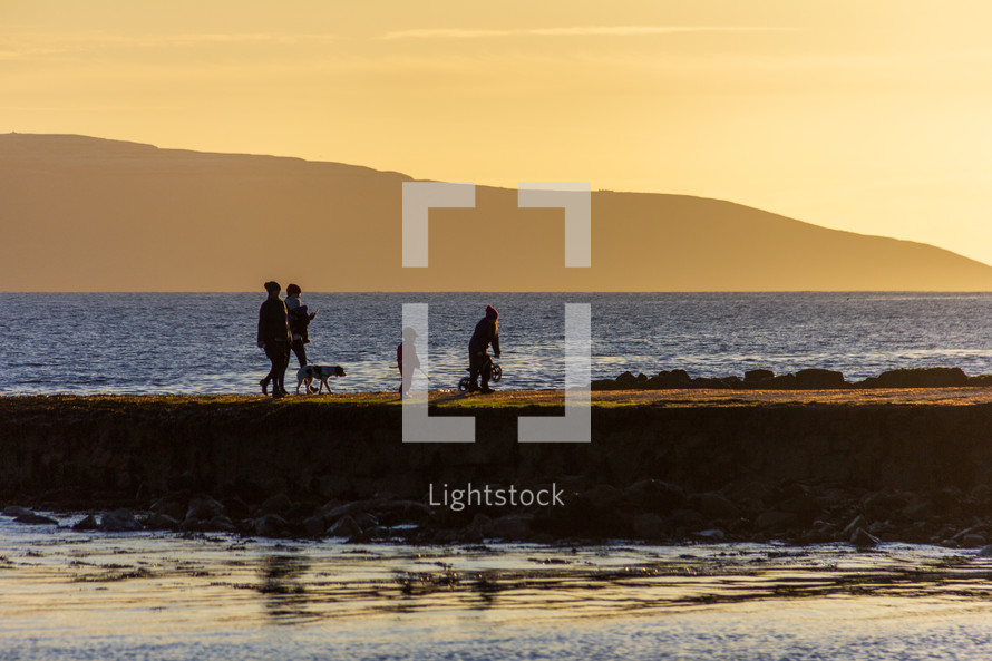 A family walking on a Salthill, Galway, Ireland jetty at sunset appear in silhouette against a yellow sky at sunset