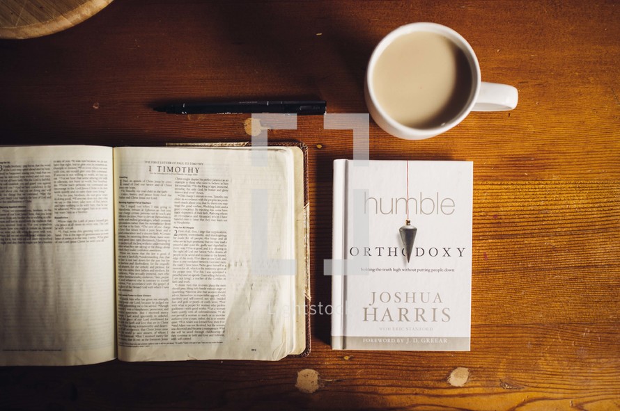 Open Bible on a wooden table with a book, pen, and a cup of coffee.