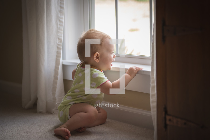 infant in a onesie looking out a window 