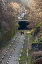 A couple walks along tree lined railroad tracks toward a tunnel