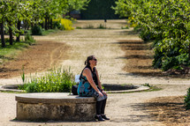 A woman sits contemplatively on a concrete bench in a grove of trees in garden near a fountain