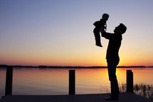 Silhouette of a father holding up his son in front of a lake as dawn breaks behind them.