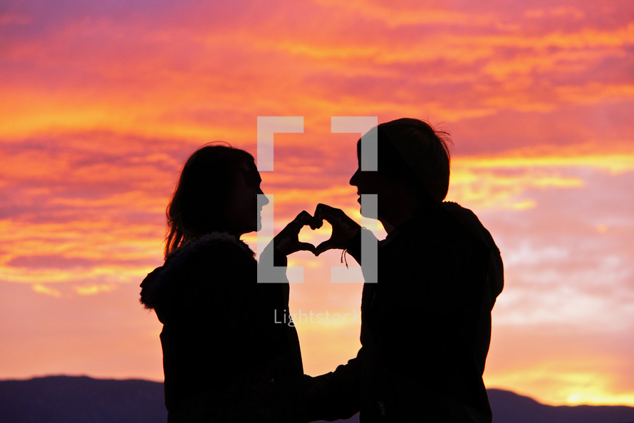 A couple make a heart shape with their hands as a symbol of their love against a backdrop of  the sunset/sunrise.