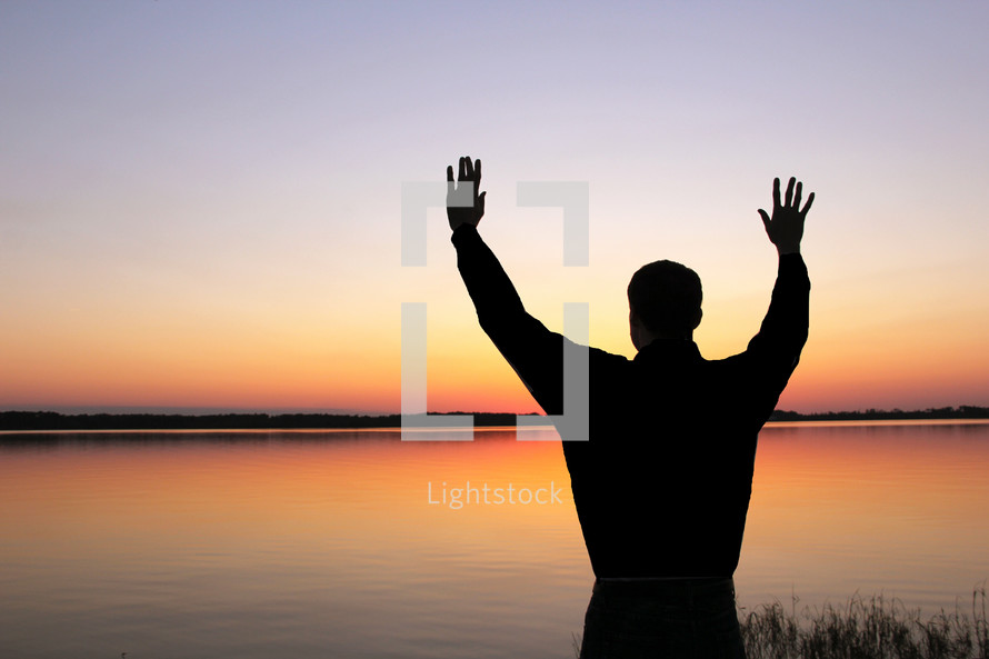 Silhouette of man with arms raised near lake at sunset.