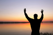 Silhouette of man with arms raised near lake at sunset.