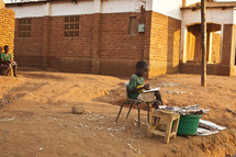 A child drawing a picture sitting in a chair outside in the dirt. 