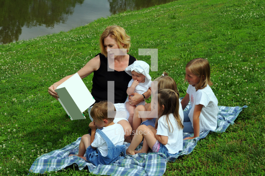 Woman reading a book to a group of children, sitting on a blanket on a hillside