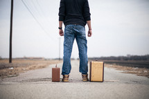 man standing looking down a road standing next to suitcases