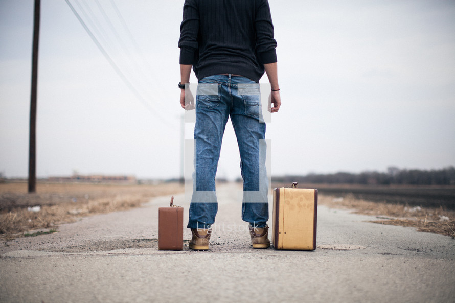man standing looking down a road standing next to suitcases