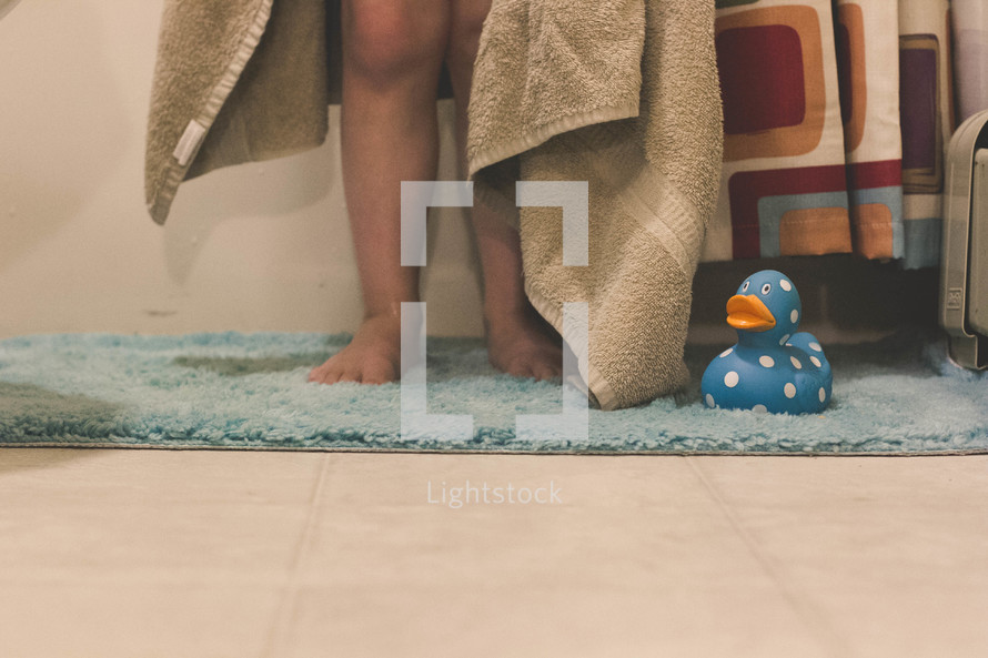 Child's legs standing on a rug with a towel and a rubber duck.