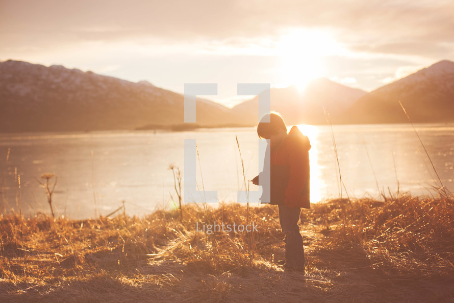 Silhouette of a boy in the sunlight near a lake surrounded by mountains.