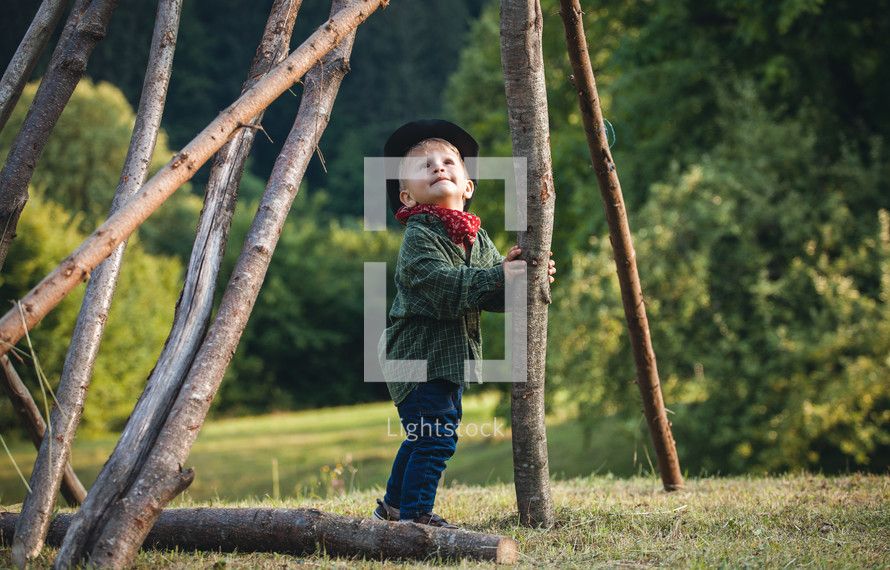 Little Cute Blond Boy in a Hat in Nature between Wooden Logs