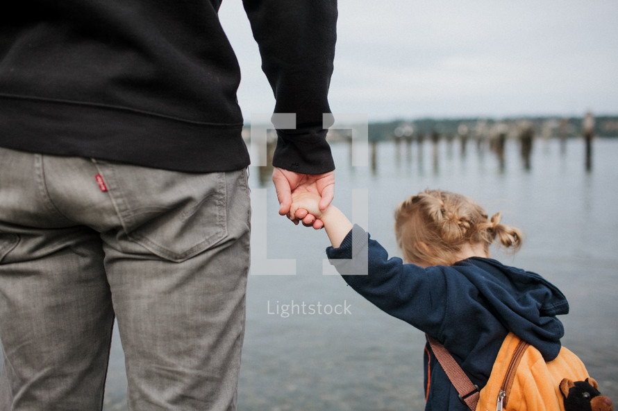 a father and toddler girl holding hands on a river shore 